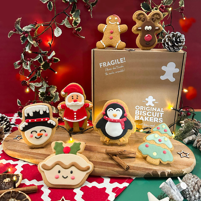 Christmas assorted party pack featuring hand-iced gingerbread and sugar cookie biscuits including Santa, reindeer, penguin, snowman, Christmas tree, gingerbread person and mince pie, displayed on a festive Christmas table.