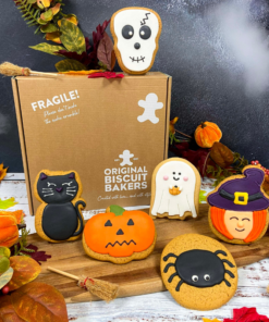 Hand-iced Halloween biscuits featuring a skull biscuit, black cat biscuit, pumpkin biscuit, ghost biscuit, spider biscuit and witch biscuit, displayed with Original Biscuit Bakers gift packaging in an autumn-inspired Halloween setting.