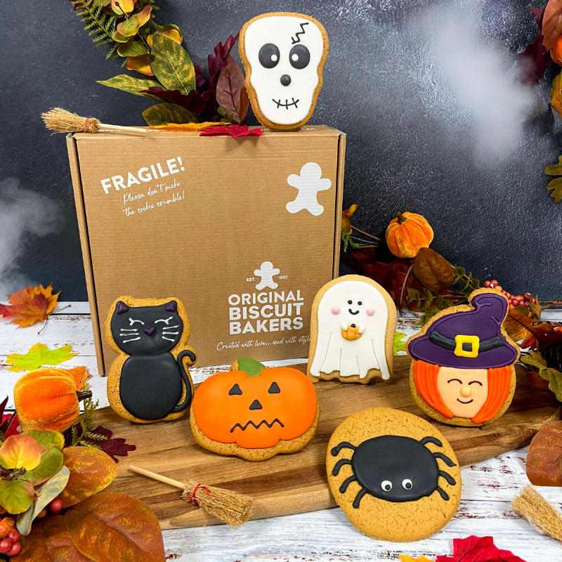 Hand-iced Halloween biscuits featuring a skull biscuit, black cat biscuit, pumpkin biscuit, ghost biscuit, spider biscuit and witch biscuit, displayed with Original Biscuit Bakers gift packaging in an autumn-inspired Halloween setting.