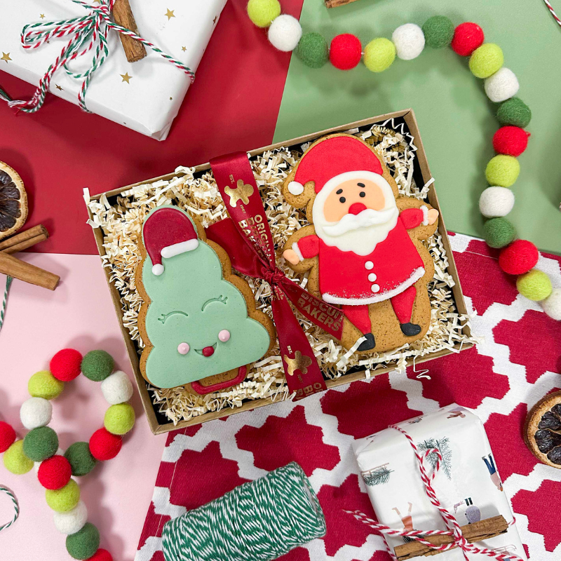 Hand-iced Christmas biscuit gift box featuring Santa and Christmas tree biscuits, presented in a festive gift tray with red ribbon and seasonal decorations.