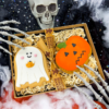 Hand-iced Halloween biscuit gift box featuring a ghost biscuit and pumpkin biscuit presented in shredded paper packaging, styled with spooky Halloween decorations including a skull, skeleton hands and cobwebs.