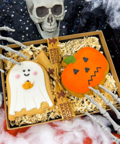 Hand-iced Halloween biscuit gift box featuring a ghost biscuit and pumpkin biscuit presented in shredded paper packaging, styled with spooky Halloween decorations including a skull, skeleton hands and cobwebs.