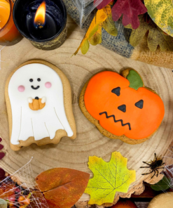 Original Biscuit Bakers hand-iced Halloween ghost and pumpkin biscuits displayed on a rustic wooden slice surrounded by autumn leaves, candles, cobweb decorations, acorns and seasonal foliage.