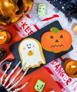 Hand-iced Halloween biscuits featuring a ghost biscuit and pumpkin biscuit displayed on a serving board within a spooky Halloween scene, surrounded by candles, skeleton hands and seasonal decorations.