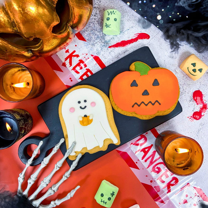 Hand-iced Halloween biscuits featuring a ghost biscuit and pumpkin biscuit displayed on a serving board within a spooky Halloween scene, surrounded by candles, skeleton hands and seasonal decorations.