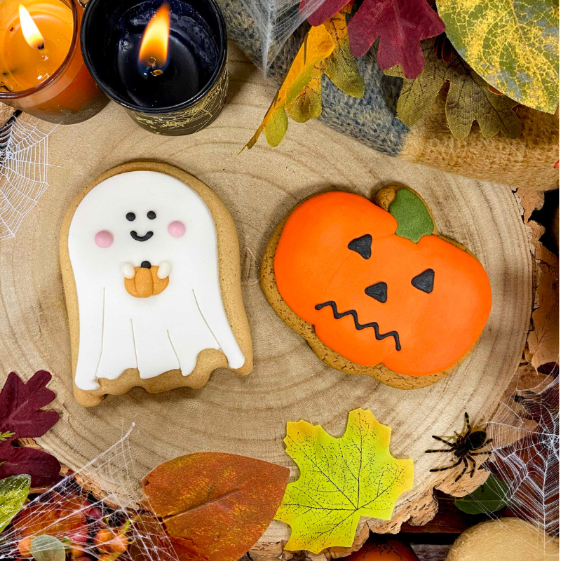 Original Biscuit Bakers hand-iced Halloween ghost and pumpkin biscuits displayed on a rustic wooden slice surrounded by autumn leaves, candles, cobweb decorations, acorns and seasonal foliage.