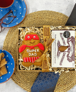 Gift box containing an Original Biscuit Bakers' “Super Dad” gingerbread biscuit and Billington’s gingerbread, presented with decorative straw on a woven placemat, styled with a mug, glasses, loose biscuits, and packaging.