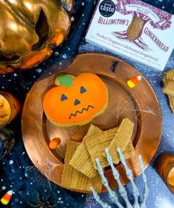Billington's Gingerbread and an Original Biscuit Bakers pumpkin biscuit displayed on a copper serving platter in a Halloween-themed setting, surrounded by candles, cobwebs, candy corn, skeleton hand decorations and seasonal spooky props.
