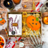 Billington's Gingerbread and an Original Biscuit Bakers pumpkin biscuit presented in a Halloween gift box with shredded paper filling, styled with spooky seasonal decorations including candles, skeleton hands, cobwebs and skull props.