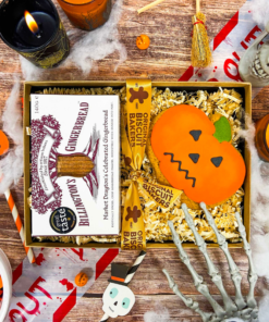 Billington's Gingerbread and an Original Biscuit Bakers pumpkin biscuit presented in a Halloween gift box with shredded paper filling, styled with spooky seasonal decorations including candles, skeleton hands, cobwebs and skull props.