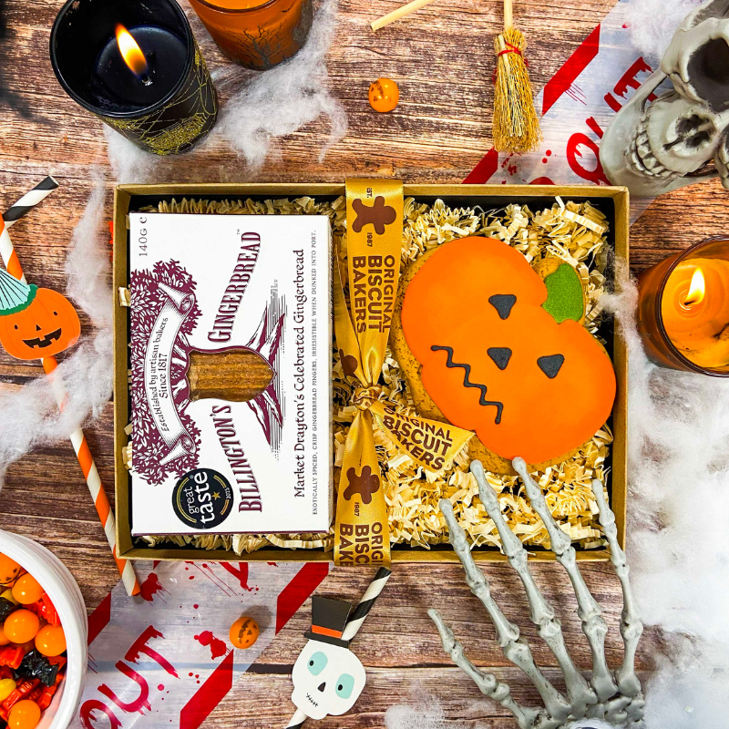 Billington's Gingerbread and an Original Biscuit Bakers pumpkin biscuit presented in a Halloween gift box with shredded paper filling, styled with spooky seasonal decorations including candles, skeleton hands, cobwebs and skull props.