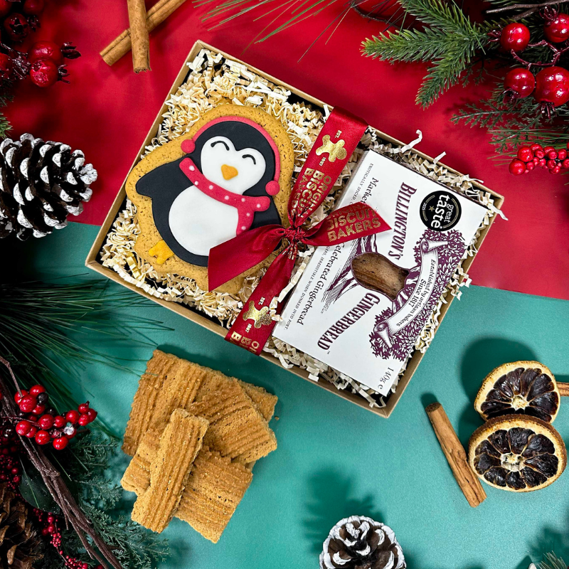 Christmas gift box featuring Billington's Gingerbread fingers and an Original Biscuit Bakers hand-iced gingerbread penguin biscuit, presented with festive ribbon and seasonal decorations.