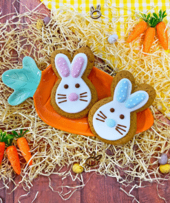 Two hand-iced gingerbread rabbit biscuits in blue and pink icing, displayed on a carrot-shaped plate surrounded by hay