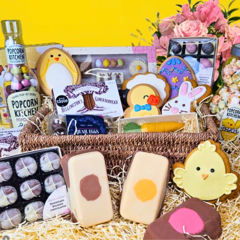Bright yellow Easter giveaway hamper filled with themed treats, including decorated iced biscuits shaped like chicks, bunnies and eggs, Billington’s gingerbread, Popcorn Kitchen popcorn bottles, assorted chocolates, fudge bars, quail‑egg sweets, and a bouquet of pink flowers arranged behind the basket. White text at the top reads ‘Easter Hamper Giveaway’.