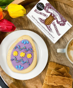 Original Biscuit Bakers Easter egg sugar cookie with colourful icing, served with Billington’s Gingerbread fingers, tulips, and a warm drink on a rustic wooden tray.