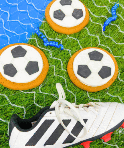 Football‑shaped iced gingerbread biscuits arranged on artificial grass with a football net and a white football boot.