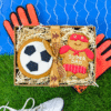 Gift box containing a football‑shaped iced gingerbread biscuit and a “Super Dad” gingerbread biscuit, presented with decorative straw, goalkeeper gloves, and football boots on artificial grass.