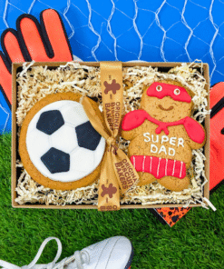 Gift box containing a football‑shaped iced gingerbread biscuit and a “Super Dad” gingerbread biscuit, presented with decorative straw, goalkeeper gloves, and football boots on artificial grass.