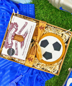 Football‑shaped iced gingerbread biscuit and Billington’s gingerbread presented in a gift box, styled with blue football boots and kit on artificial grass.