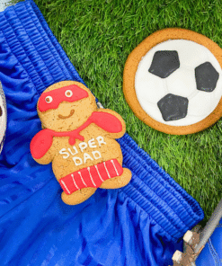 Football‑shaped iced gingerbread biscuit and a “Super Dad” gingerbread biscuit displayed on artificial grass with blue football shorts, a football, and football boots.
