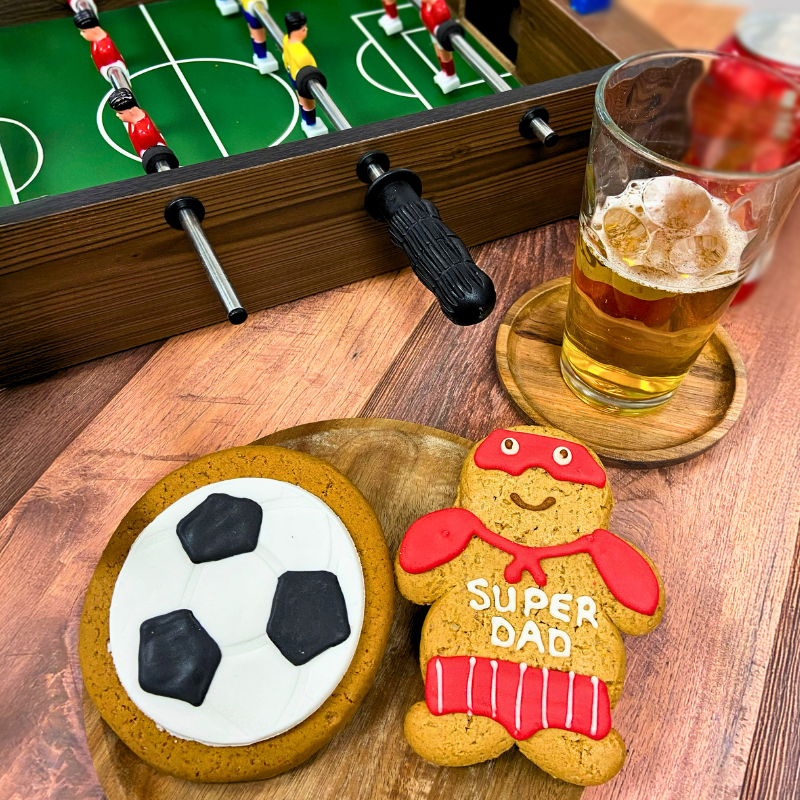 Football themed gingerbread biscuits featuring an iced football biscuit and a Super Dad biscuit, styled with table football and a drink.