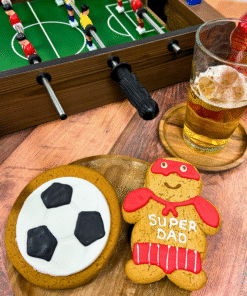 Football themed gingerbread biscuits featuring an iced football biscuit and a Super Dad biscuit, styled with table football and a drink.