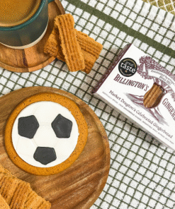 Football‑shaped iced gingerbread biscuit on a wooden board, styled with Billington’s gingerbread, loose gingerbread biscuits, and a mug on a checked kitchen cloth.