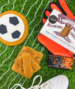 Football‑shaped iced gingerbread biscuit displayed with Billington’s gingerbread, goalkeeper gloves, and football boots on artificial grass with goal net.