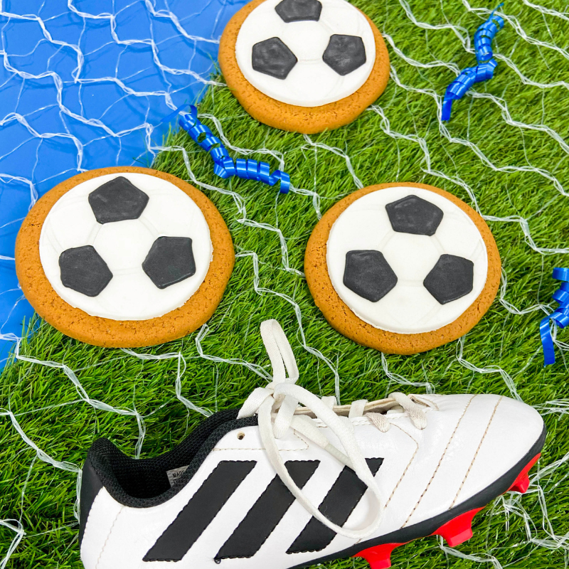 Hand‑iced football gingerbread biscuits by Original Biscuit Bakers arranged on grass turf with a football boot and net, ideal for sports events, party bags, and gift shops.