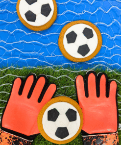 Football‑shaped iced gingerbread biscuits positioned in front of a goal net on artificial grass with bright orange goalkeeper gloves.