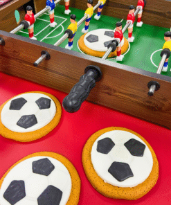 Football‑shaped iced gingerbread biscuits displayed beside a tabletop football game with wooden players and metal rods on a red and green surface.