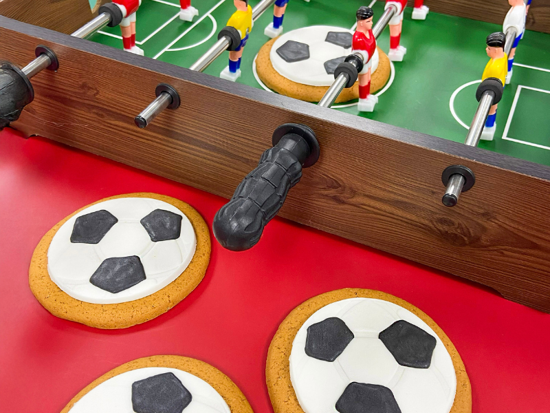 Football‑shaped iced gingerbread biscuits displayed beside a tabletop football game with wooden players and metal rods on a red and green surface.