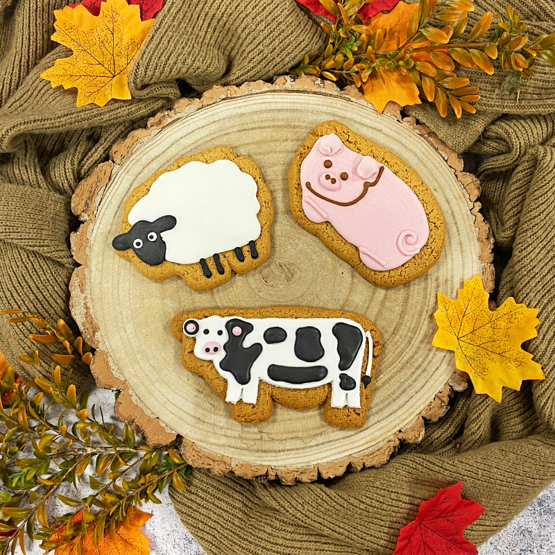 Hand-iced farm animal gingerbread biscuits featuring a sheep, pig and cow, styled on a wooden slice with autumn leaves and cosy knitwear.