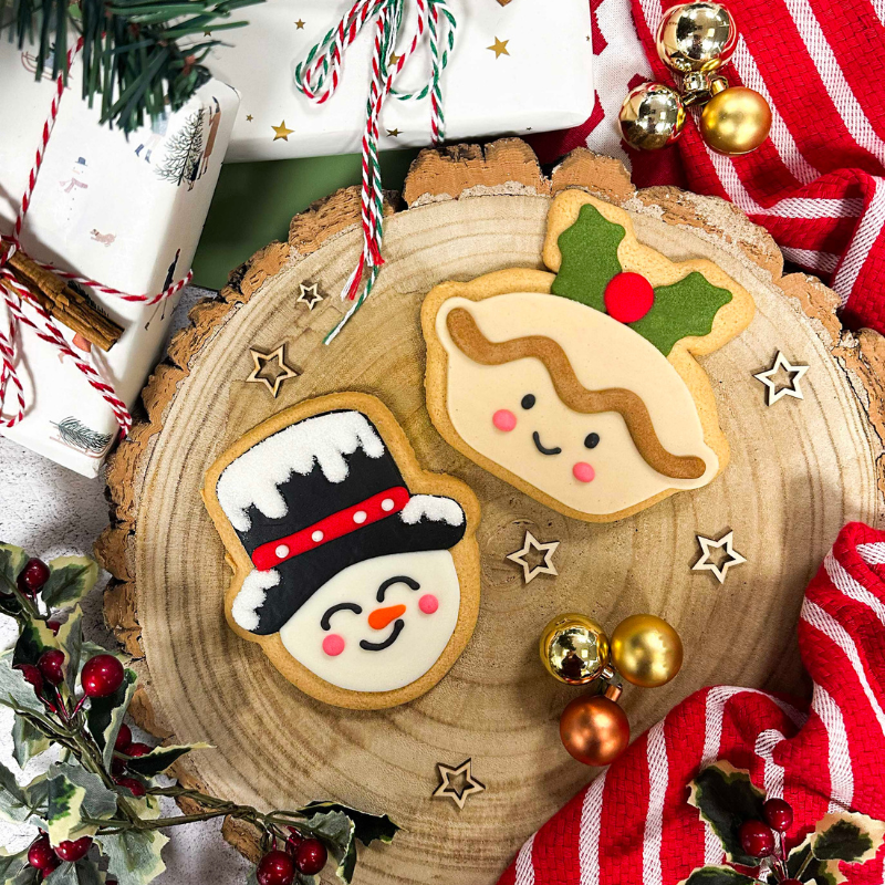 Hand-iced sugar cookie mince pie and snowman biscuits displayed on a rustic wooden serving board with festive Christmas decorations.
